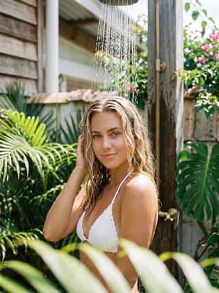 Blonde with sun-kissed waves smiling under a tropical outdoor shower surrounded by lush palm fronds