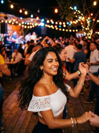 Curly-haired Latina laughing while dancing at an outdoor string-lit patio party in a white lace crop top