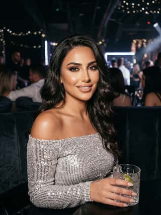 Dark-haired woman in a silver off-shoulder sequin top holding a cocktail in a crowded dimly lit nightclub