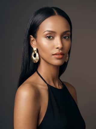 Sleek black-haired woman with gold hoop earrings in a black halter top, dramatic studio lighting on a dark backdrop