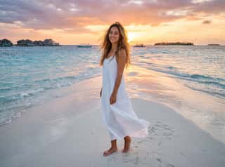 Barefoot brunette in a flowing white maxi dress strolling along the shore at sunset with overwater villas behind