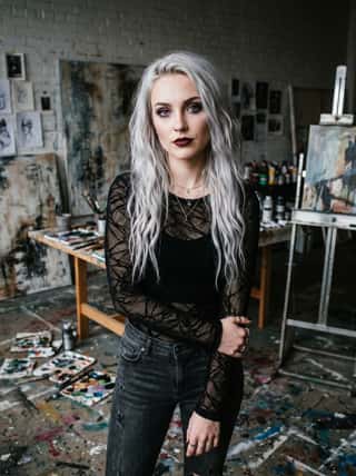 Silver-haired woman with dark lipstick in a black mesh top standing in a paint-splattered artist studio