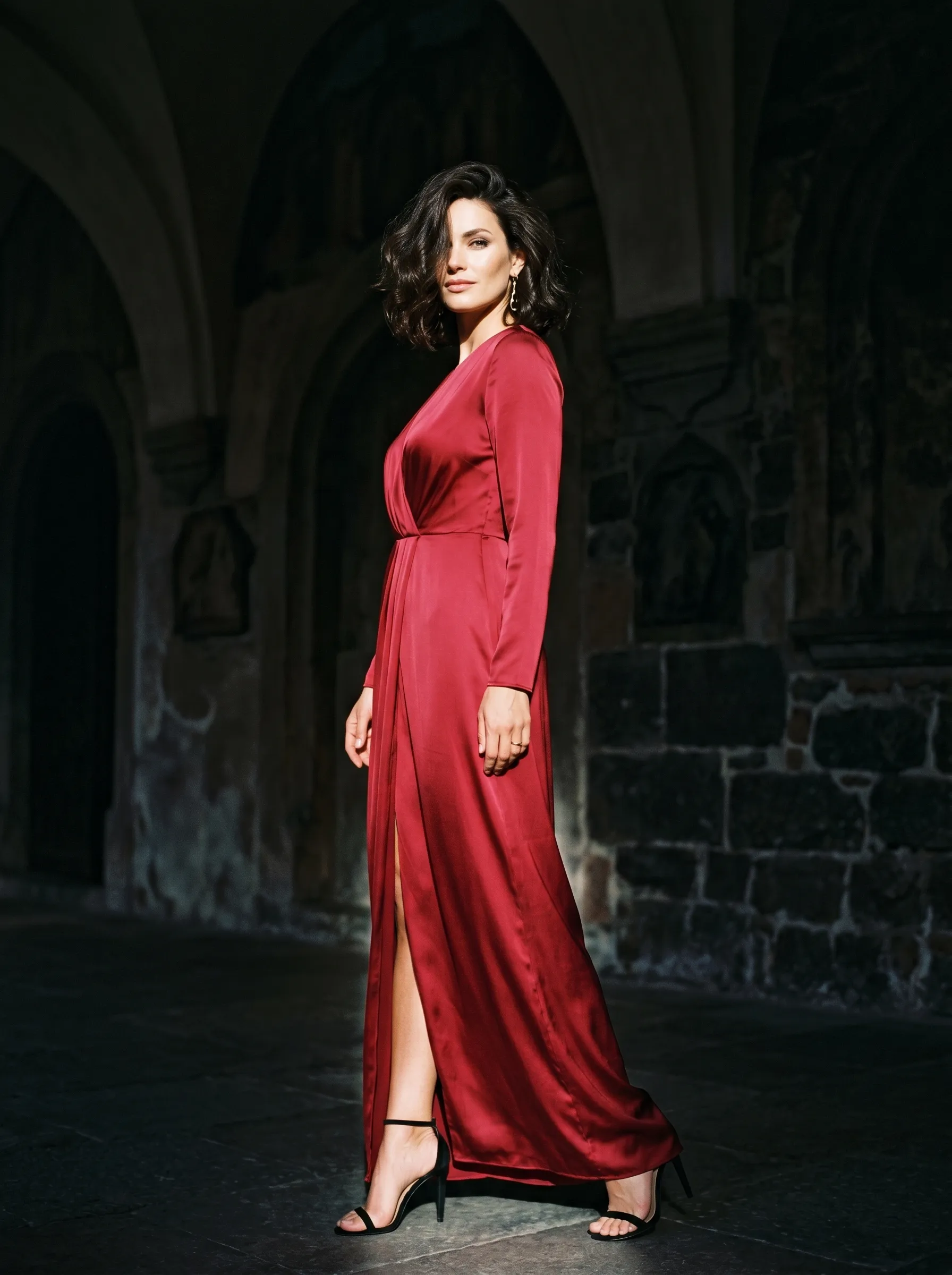 Brunette with wavy bob in a flowing red satin gown, standing beneath stone arches with dramatic side lighting